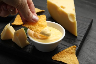 Photo of Woman dipping nacho chip into tasty cheese sauce at black table, closeup