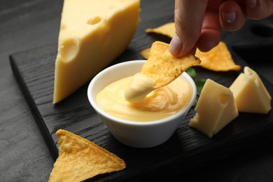 Photo of Woman dipping nacho chip into tasty cheese sauce at black table, closeup