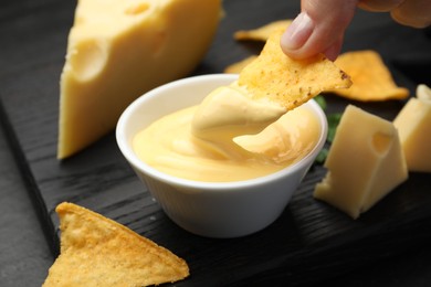 Photo of Woman dipping nacho chip into tasty cheese sauce at table, closeup