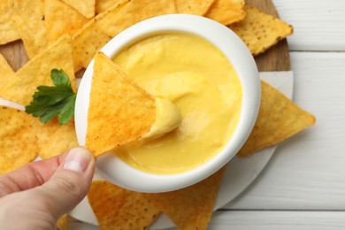 Photo of Woman dipping nacho chip into tasty cheese sauce at white wooden table, top view