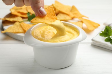 Photo of Woman dipping nacho chip into tasty cheese sauce at white wooden table, closeup