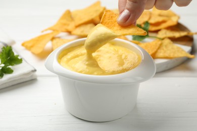 Photo of Woman dipping nacho chip into tasty cheese sauce at white wooden table, closeup