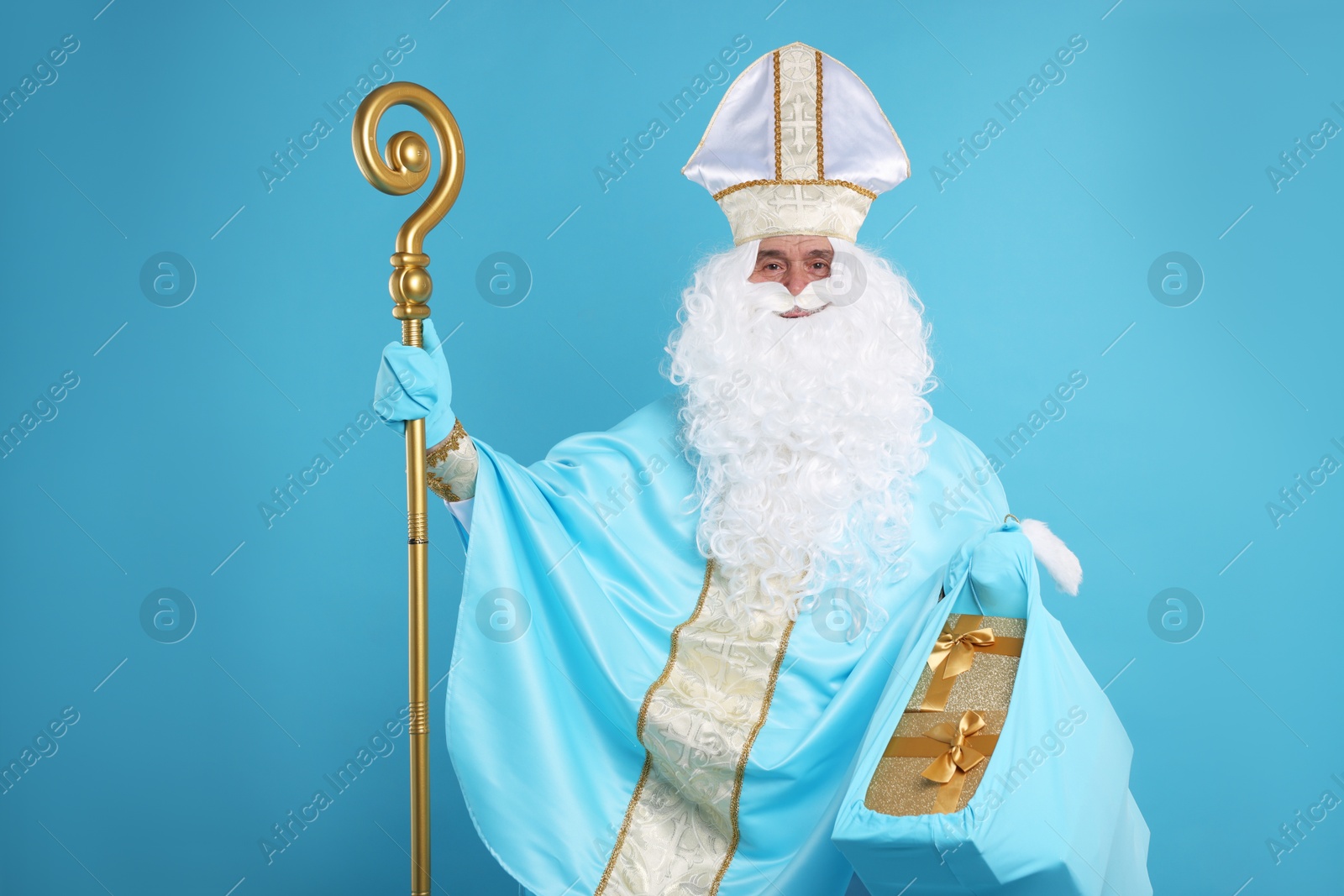 Photo of Saint Nicholas with bag of Christmas gifts and crozier on light blue background