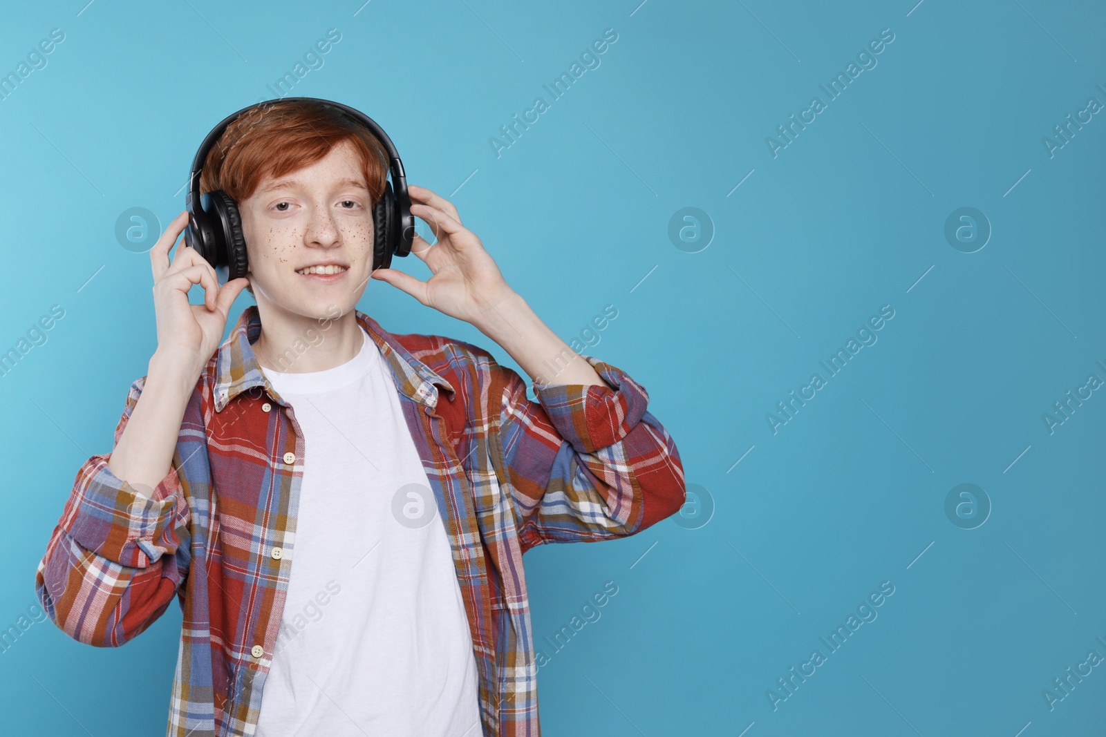 Photo of Smiling teenage boy with freckles listening to music by headphones on light blue background. Space for text