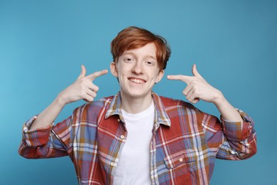 Photo of Smiling teenage boy pointing at his freckles on light blue background