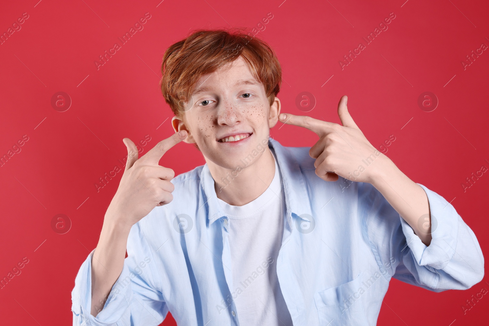 Photo of Smiling teenage boy pointing at his freckles on red background