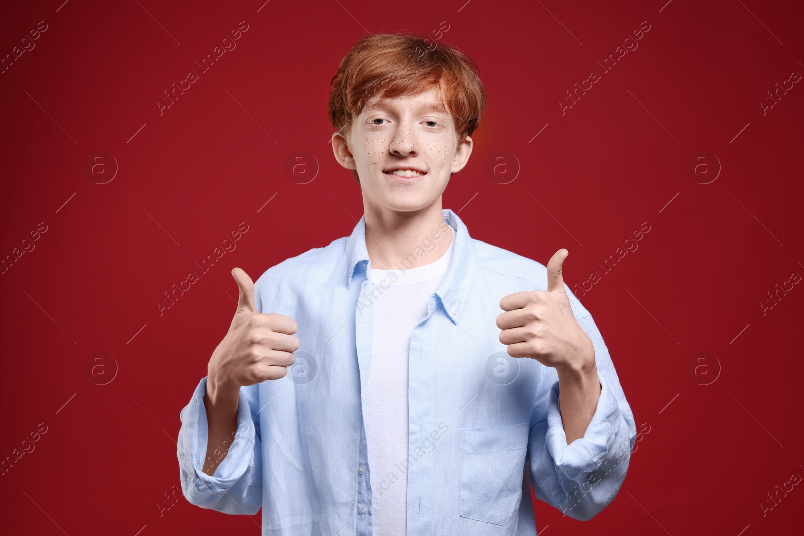 Photo of Smiling teenage boy with freckles showing thumbs up on dark red background