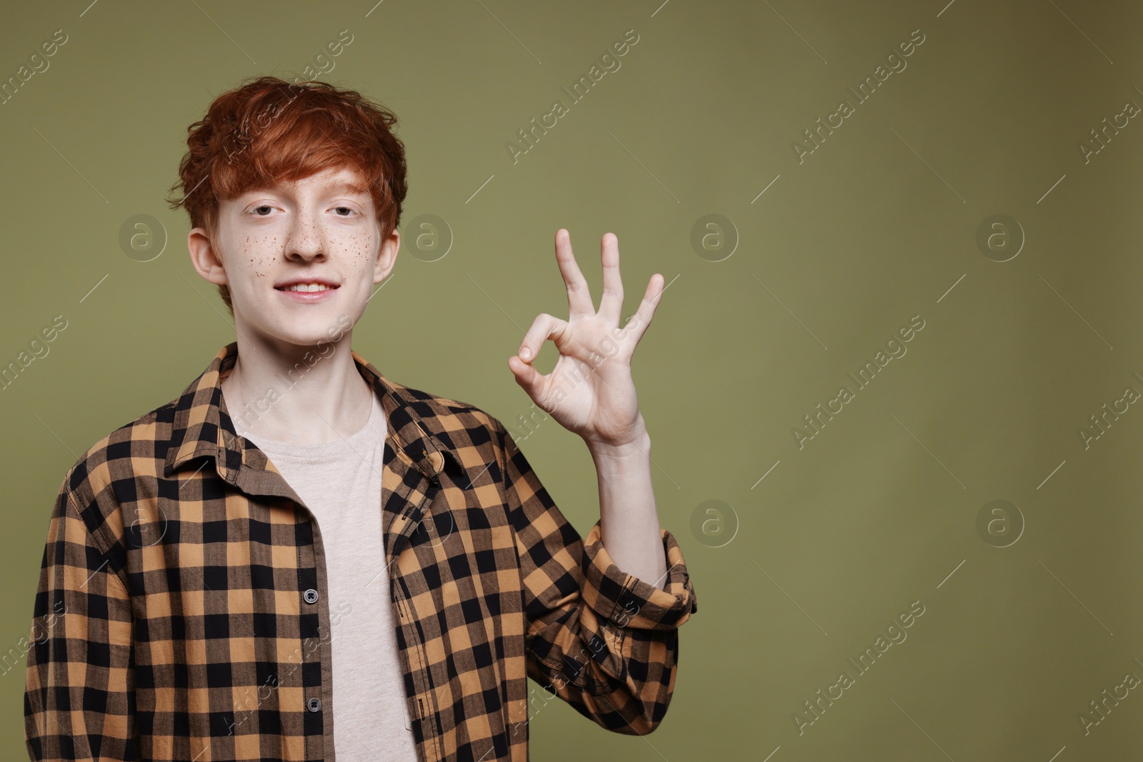 Photo of Smiling teenage boy with freckles showing ok gesture on dark green background. Space for text