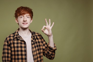 Smiling teenage boy with freckles showing ok gesture on dark green background. Space for text