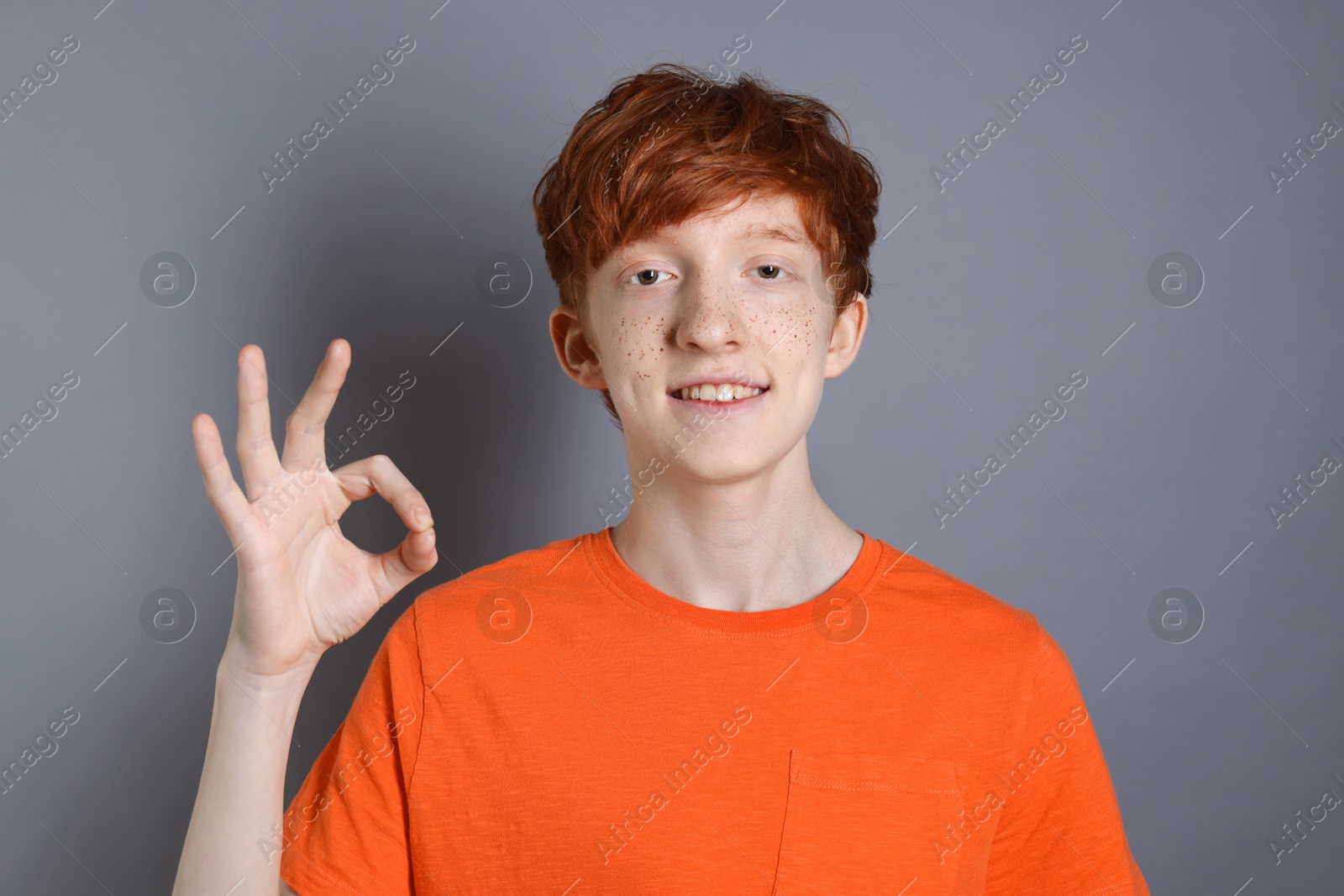 Photo of Smiling teenage boy with freckles pointing ok gesture on grey background