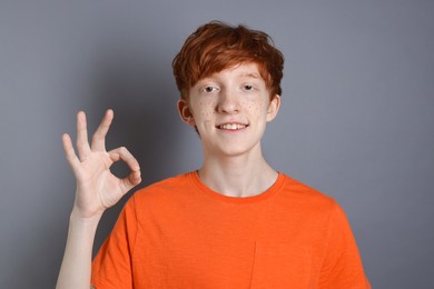 Smiling teenage boy with freckles pointing ok gesture on grey background