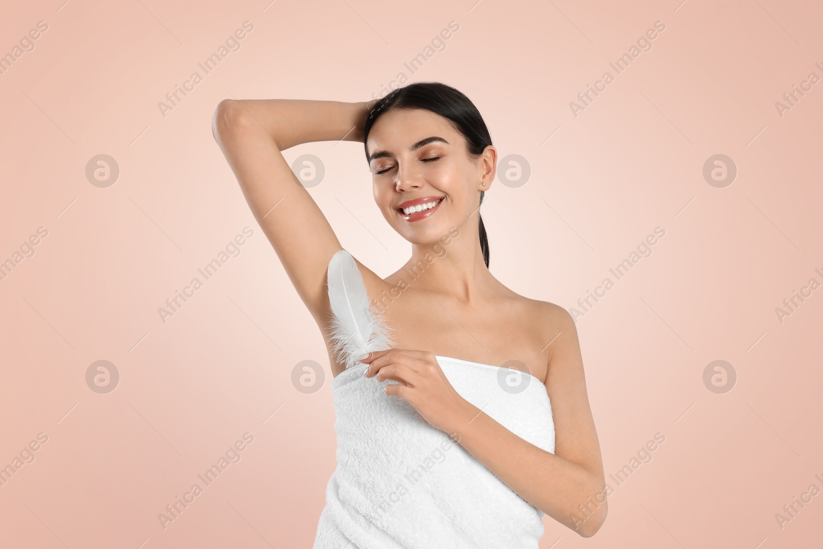 Image of Young woman touching hairless armpit with feather after epilation procedure on peach color background