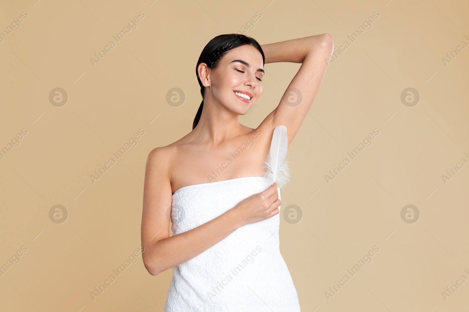 Image of Young woman touching hairless armpit with feather after epilation procedure on dark beige background