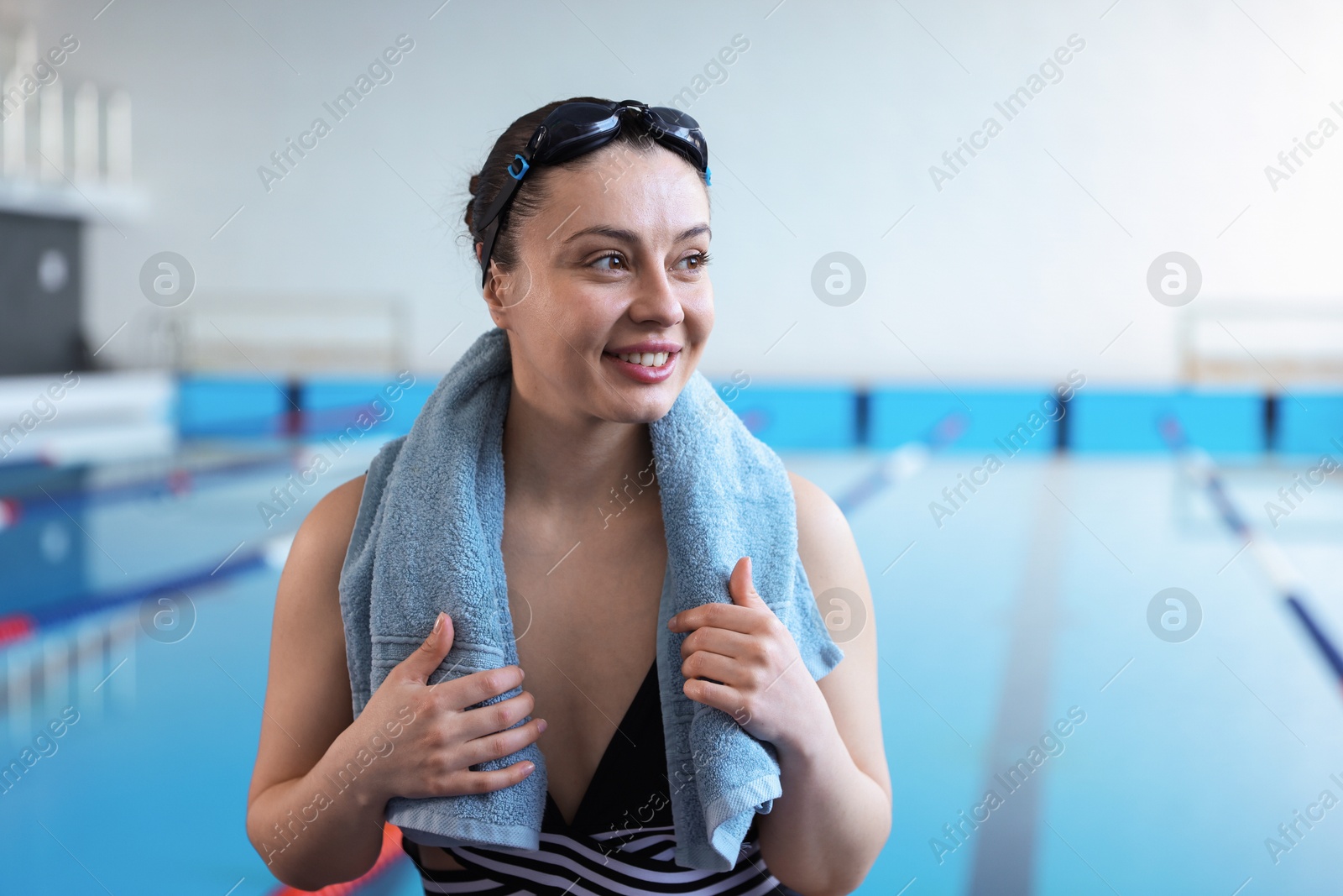 Photo of Happy woman wearing goggles and swimsuit with towel near swimming pool indoors, space for text