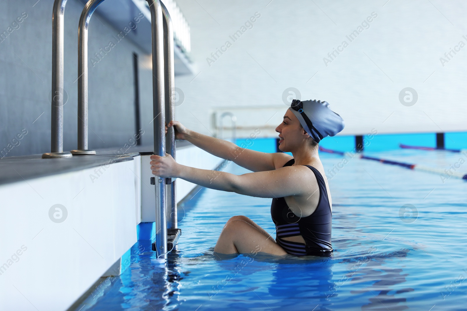 Photo of Happy woman in cap, goggles and swimsuit getting out of swimming pool indoors