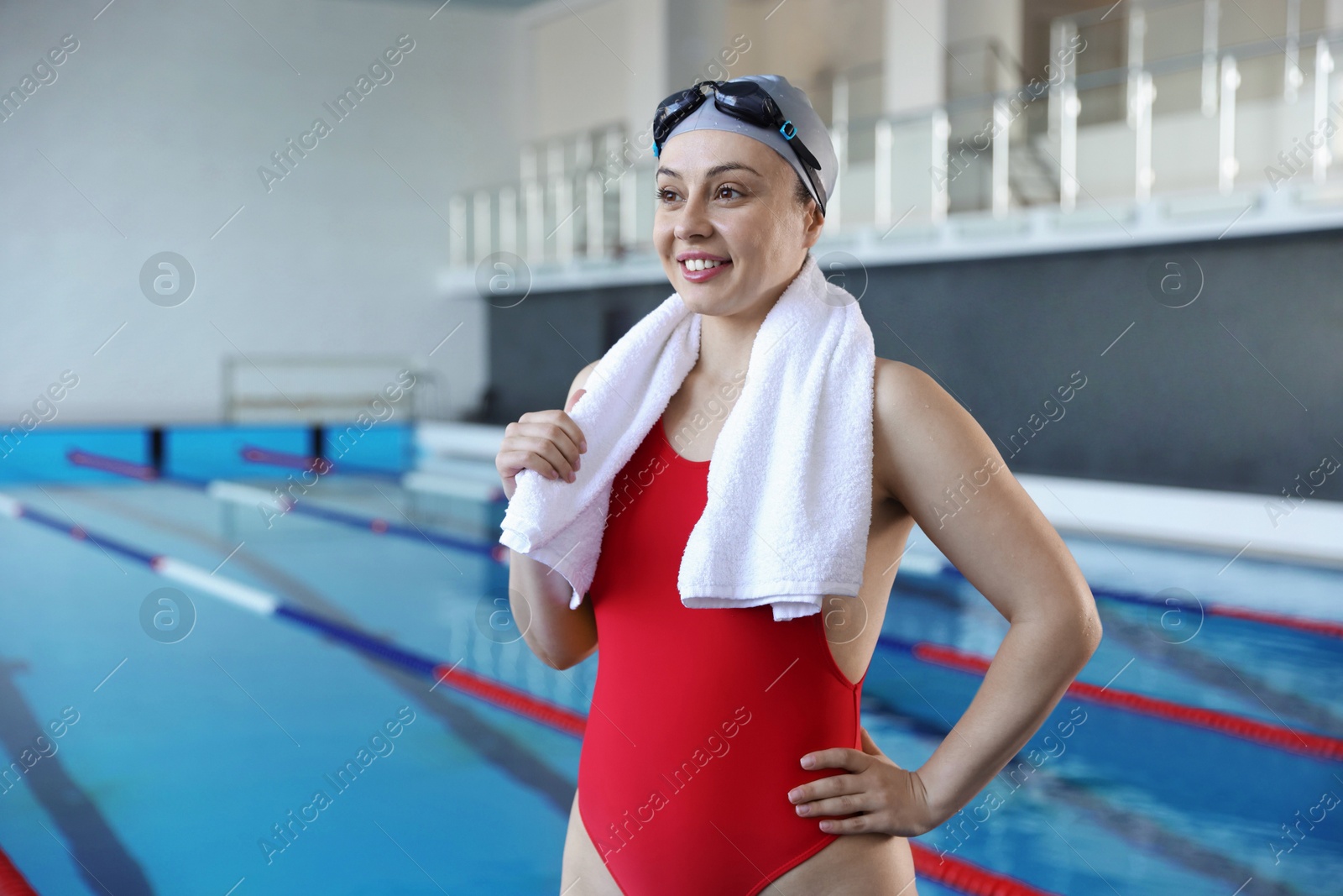 Photo of Happy woman in cap, goggles and swimsuit with towel near swimming pool indoors, space for text