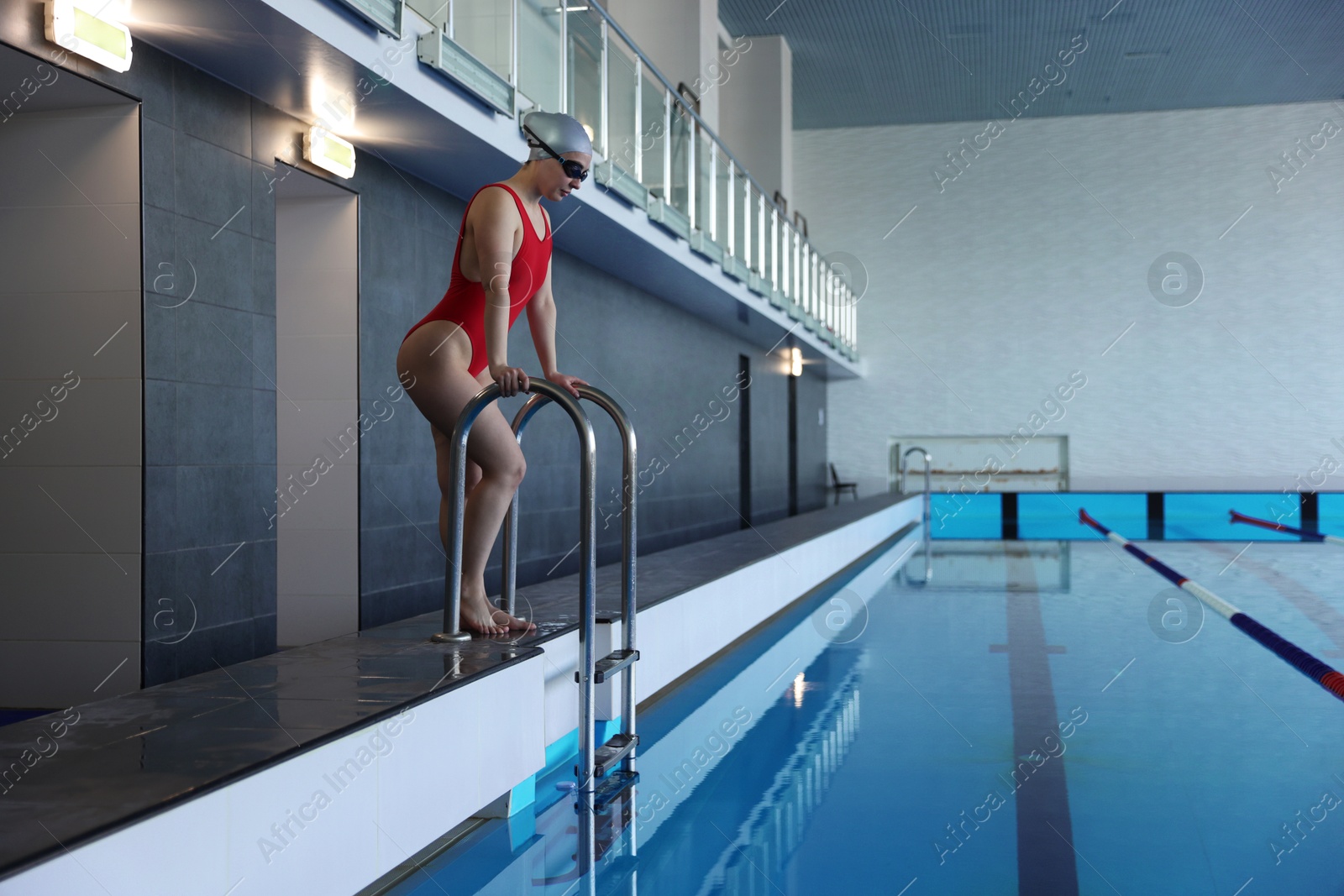 Photo of Woman wearing cap, goggles and swimsuit near swimming pool indoors, space for text