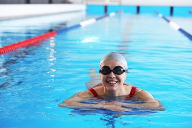 Happy woman in cap and goggles with kickboard swimming in pool indoors