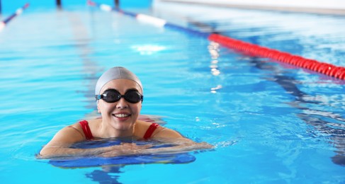 Happy woman in cap and goggles with kickboard swimming in pool indoors, space for text