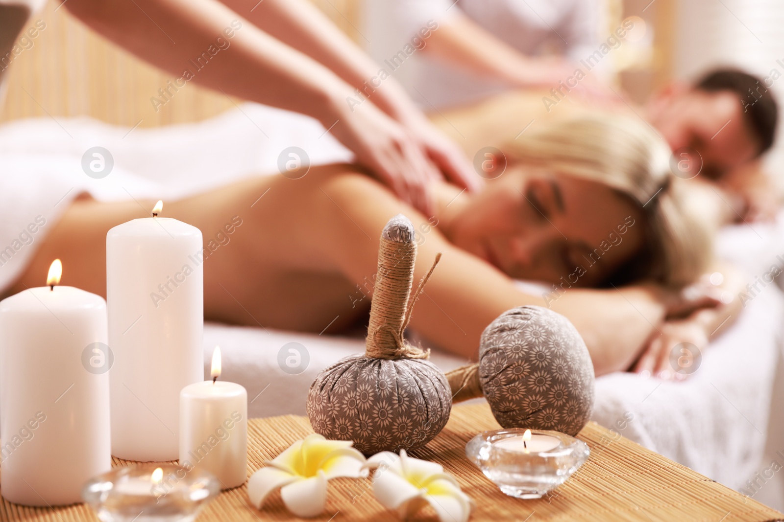 Photo of Couple receiving relaxing massage in spa salon, focus on burning candles, flowers and herbal bags