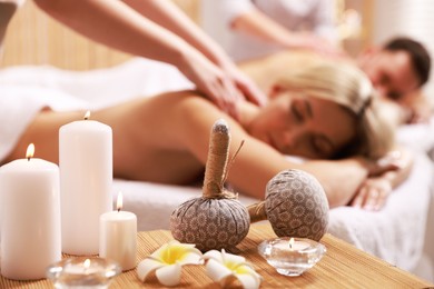 Couple receiving relaxing massage in spa salon, focus on burning candles, flowers and herbal bags