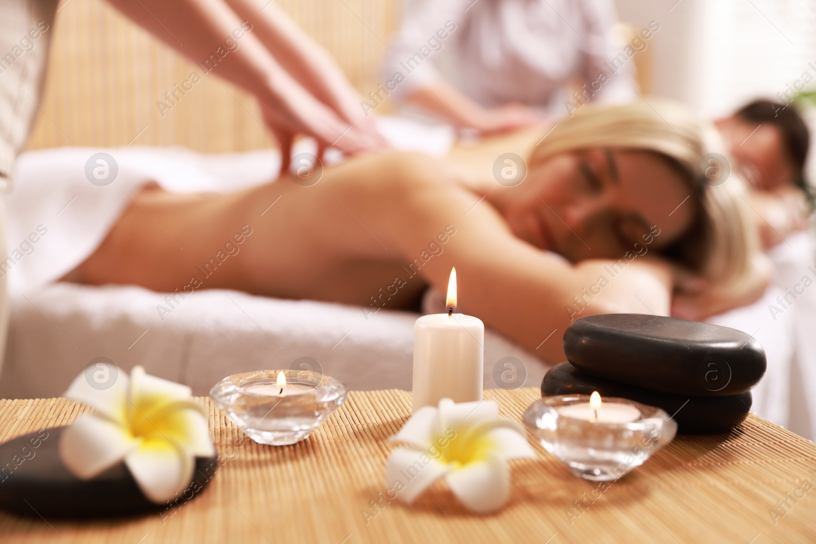 Photo of Couple receiving relaxing massage in spa salon, focus on burning candles, flowers and stones