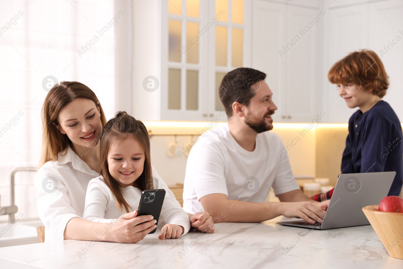 Photo of Happy family wearing stylish pajamas looking at devices at white marble table in kitchen