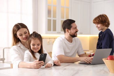 Photo of Happy family wearing stylish pajamas looking at devices at white marble table in kitchen