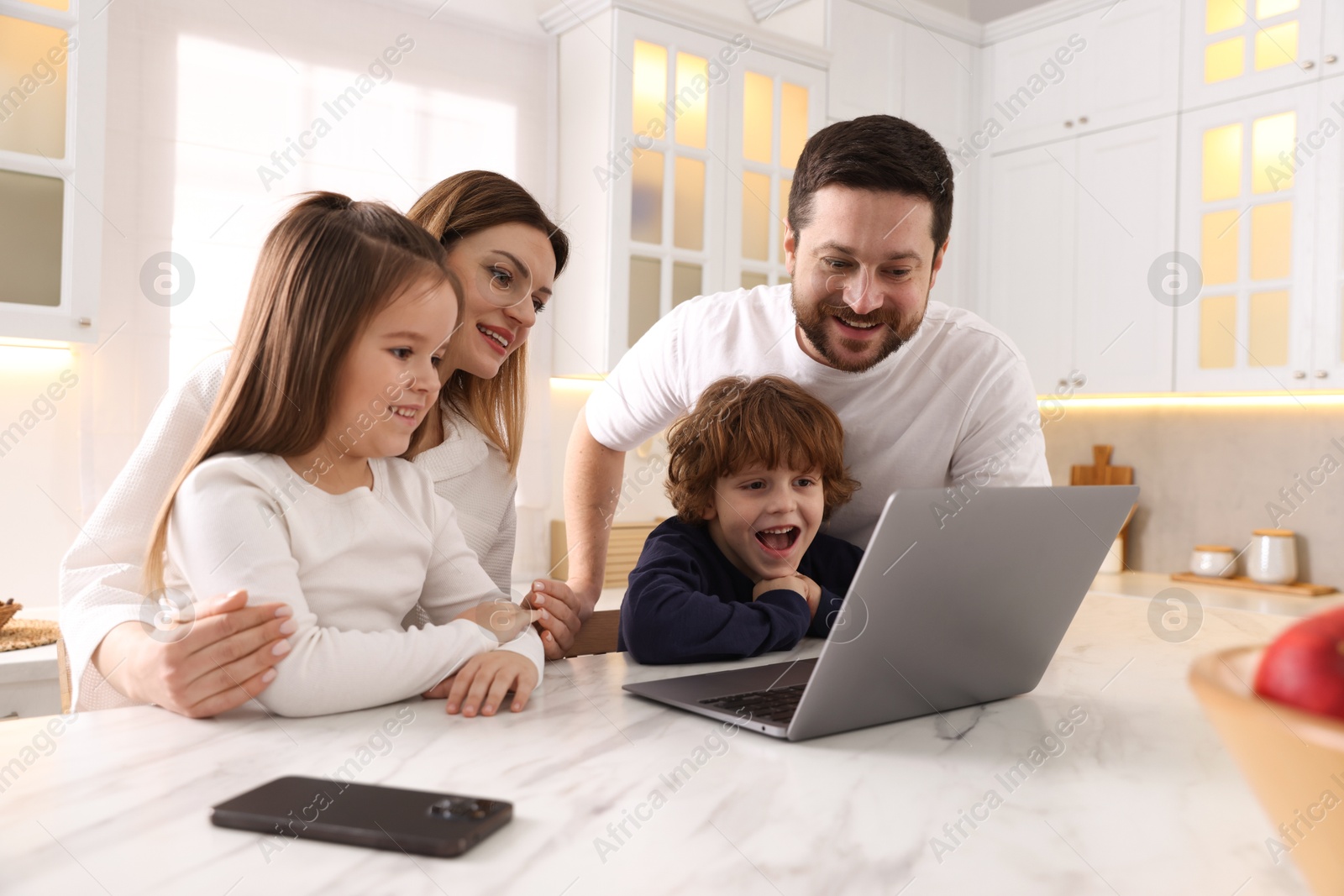 Photo of Happy family wearing stylish pajamas looking at laptop at white marble table in kitchen