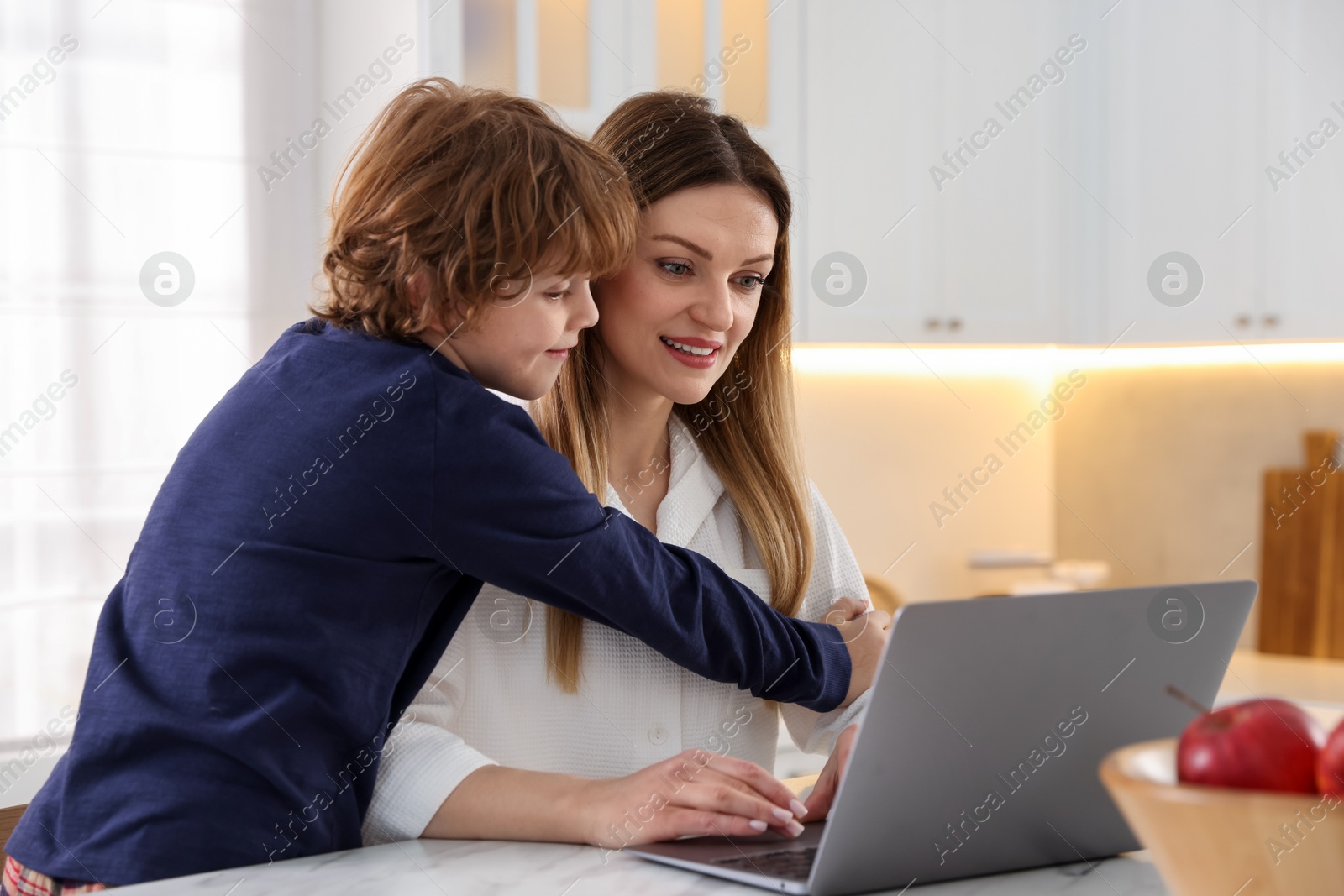 Photo of Smiling woman and her son wearing stylish pajamas with laptop at table in kitchen