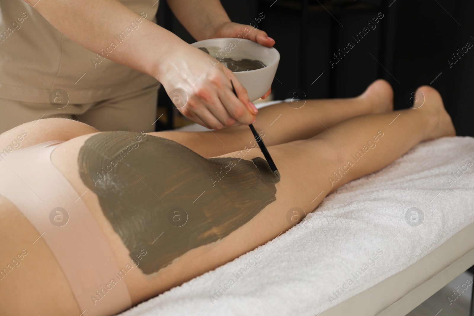 Photo of Esthetician applying cosmetic product for body wraps treatment onto woman's buttocks in spa salon, closeup