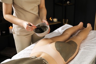 Photo of Esthetician applying cosmetic product for body wraps treatment onto woman's buttocks in spa salon, closeup