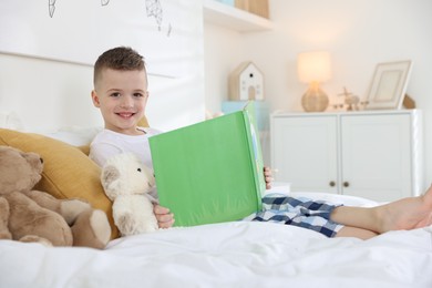 Photo of Bedtime. Cute boy reading book to his toys on bed indoors