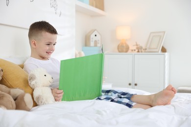 Photo of Bedtime. Cute boy reading book to his toys on bed indoors