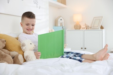 Photo of Bedtime. Cute boy reading book to his toys on bed indoors