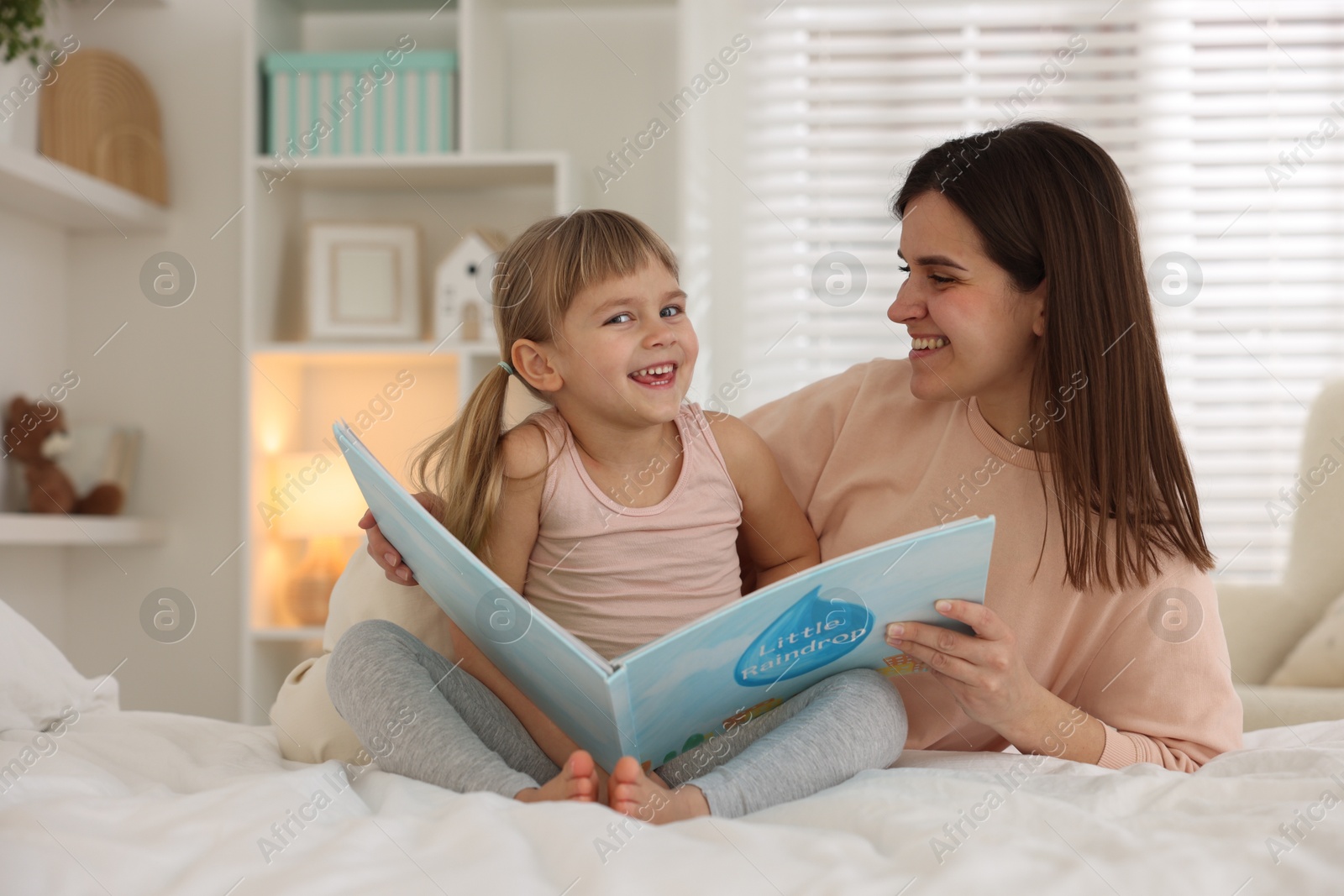 Photo of Bedtime. Happy little daughter reading book with her mother on bed at home