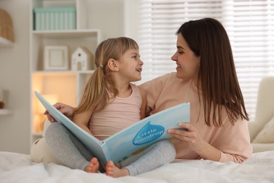 Photo of Bedtime. Happy little daughter reading book with her mother on bed at home