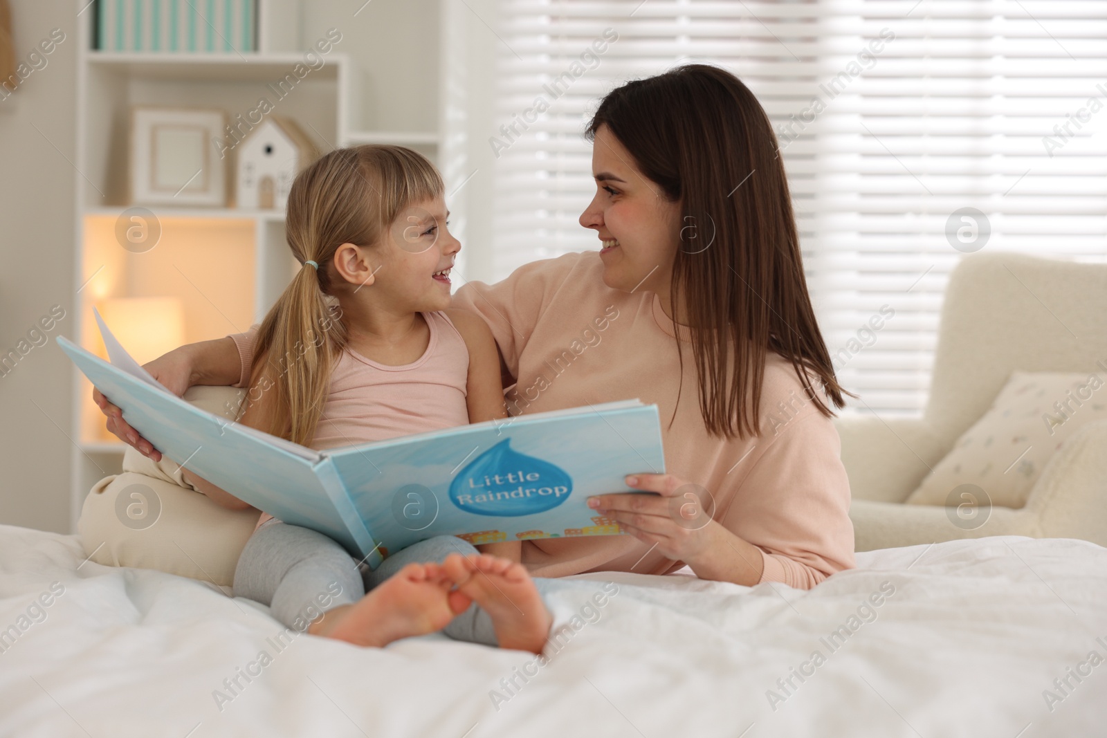 Photo of Bedtime. Happy little daughter reading book with her mother on bed at home