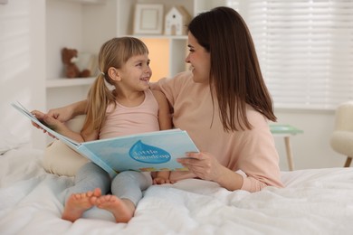 Bedtime. Happy little daughter reading book with her mother on bed at home