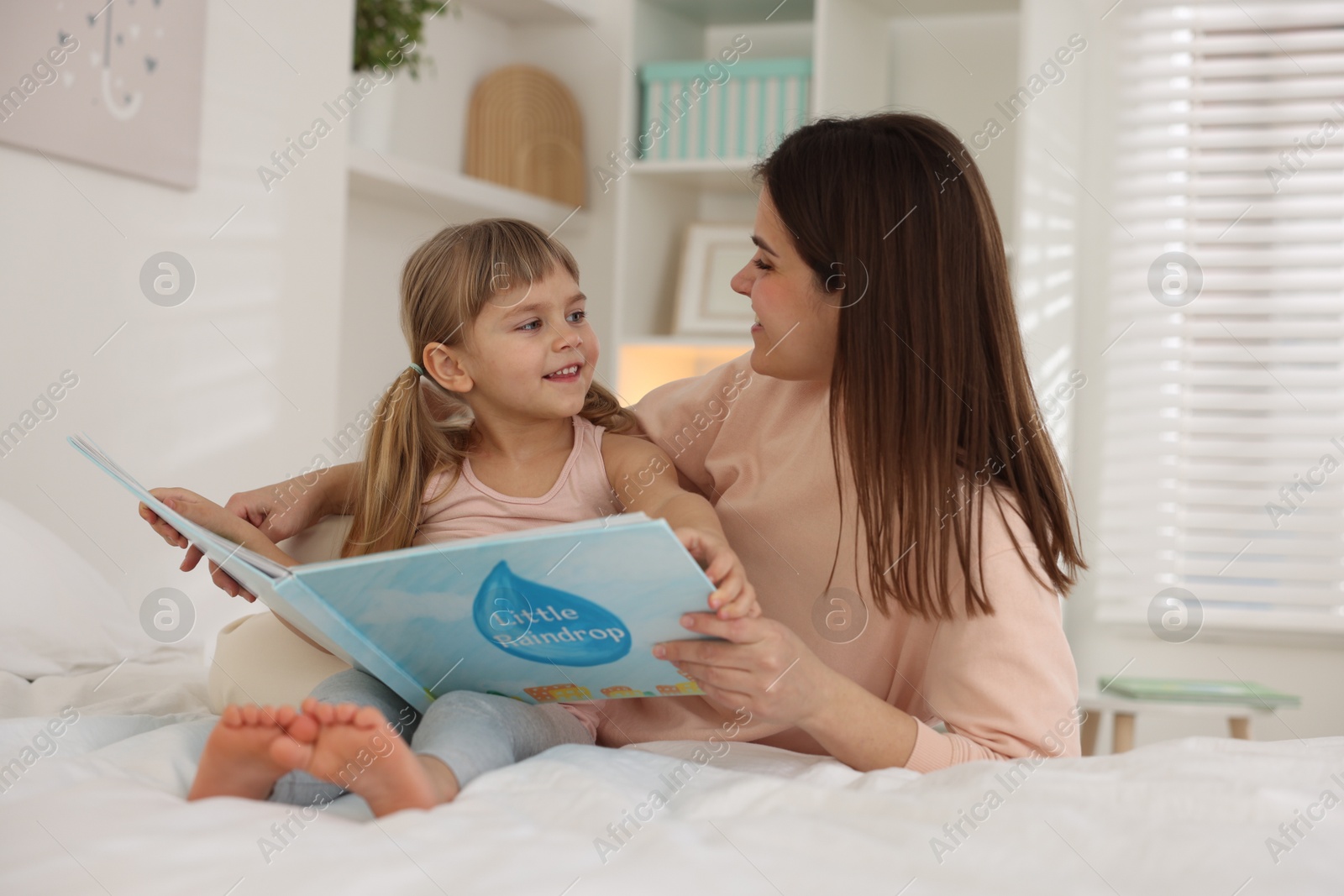 Photo of Bedtime. Happy little daughter reading book with her mother on bed at home