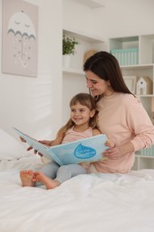 Bedtime. Happy little daughter reading book with her mother on bed at home