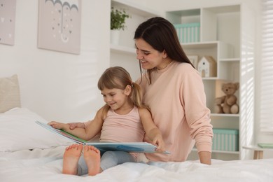 Photo of Bedtime. Happy little daughter reading book with her mother on bed at home