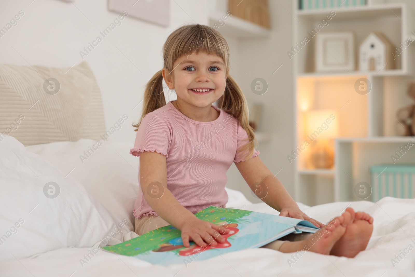 Photo of Bedtime. Happy little girl with book sitting on bed at home