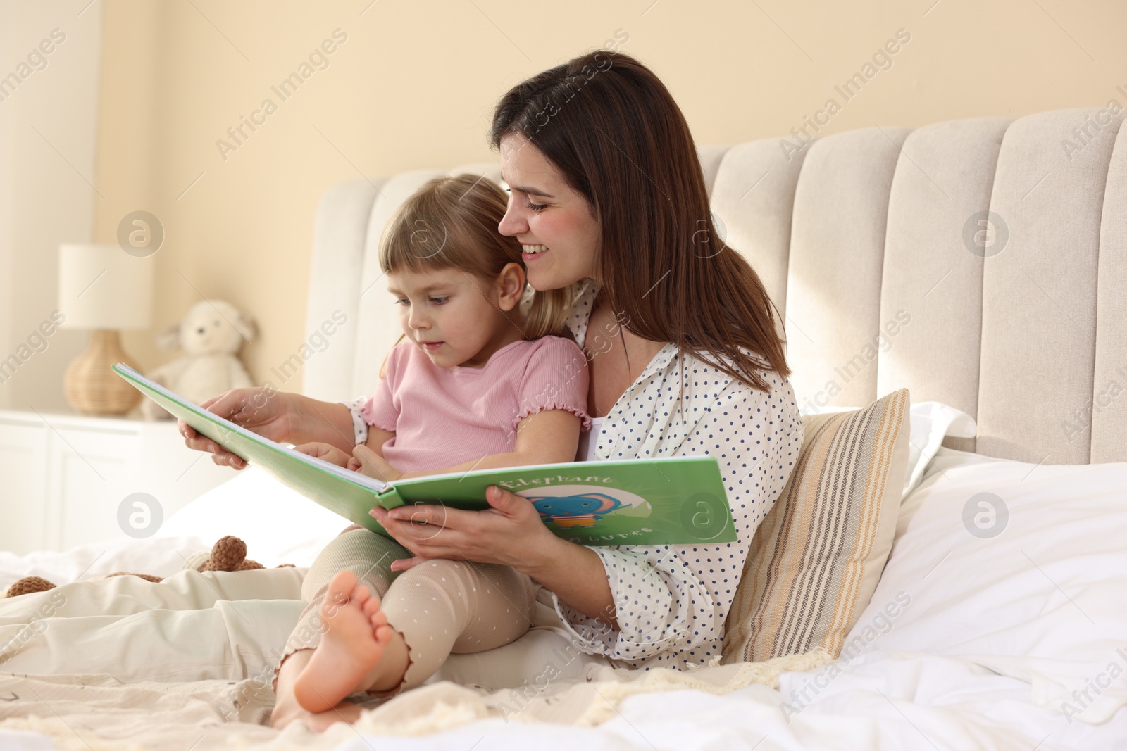 Photo of Bedtime. Cute little daughter reading book with her mother on bed at home