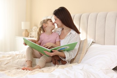 Photo of Bedtime. Happy little daughter reading book with her mother on bed at home