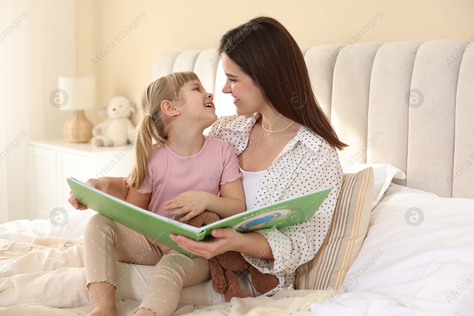 Photo of Bedtime. Happy little daughter reading book with her mother on bed at home