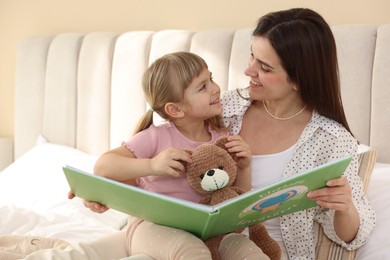 Bedtime. Happy little daughter reading book with her mother on bed at home