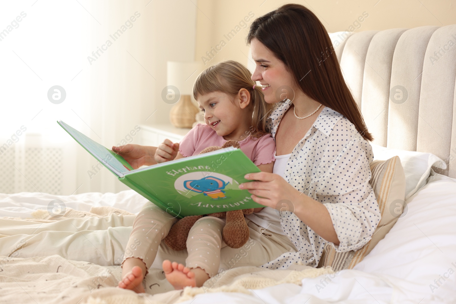 Photo of Bedtime. Happy little daughter reading book with her mother on bed at home