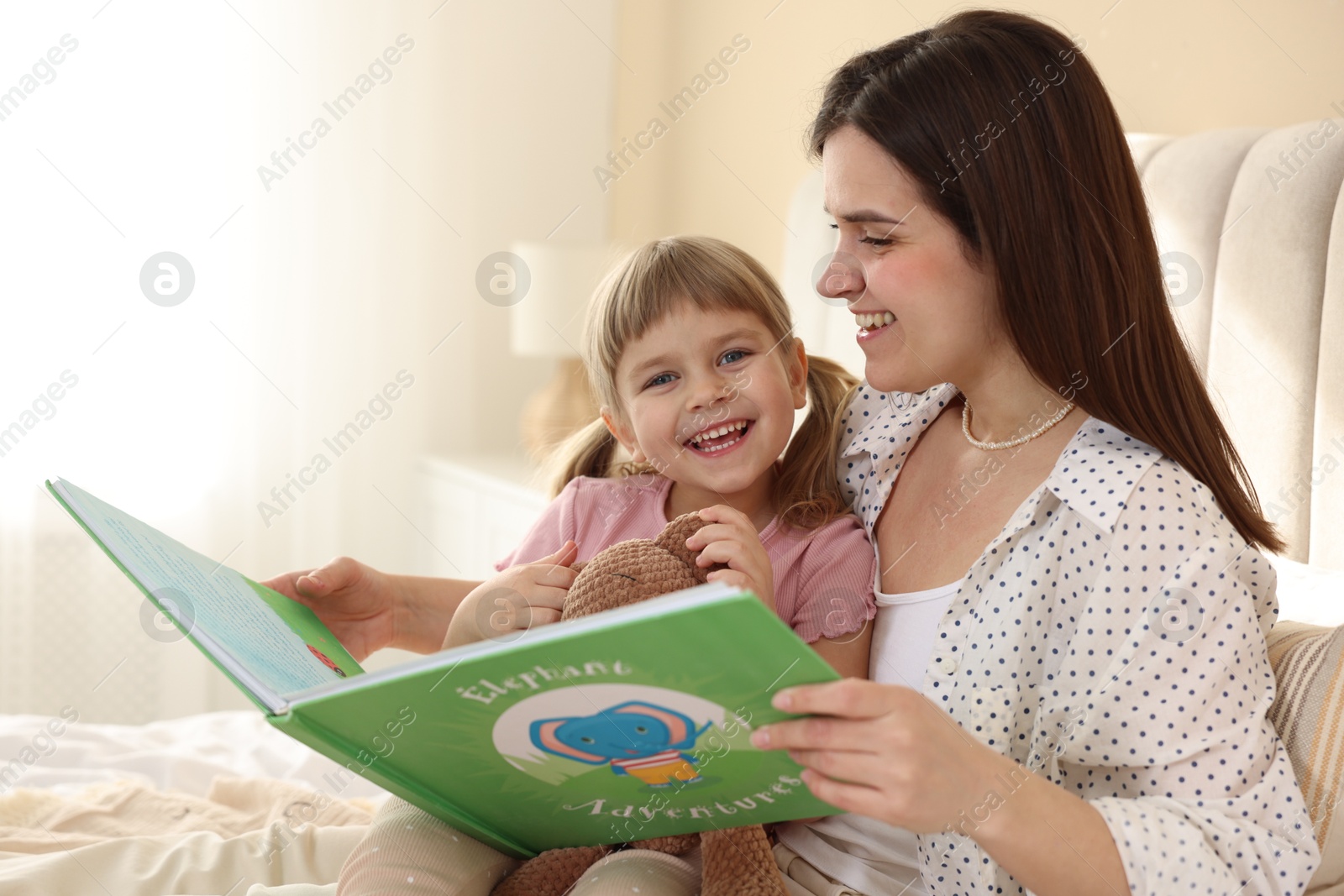 Photo of Bedtime. Happy little daughter reading book with her mother on bed at home