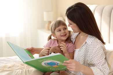 Photo of Bedtime. Happy little daughter reading book with her mother on bed at home